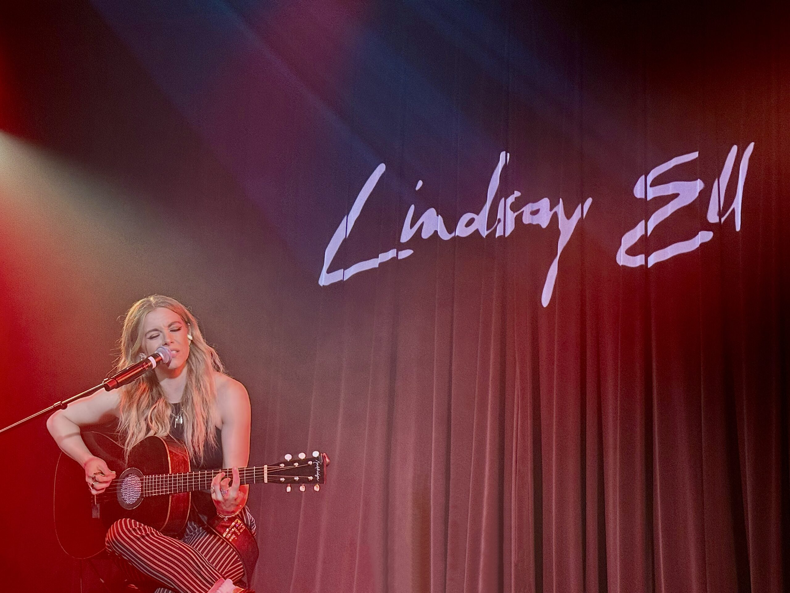 Singer Lindsay Ell performing on stage in Toronto sitting in front of a Lindsay Ell backdrop.