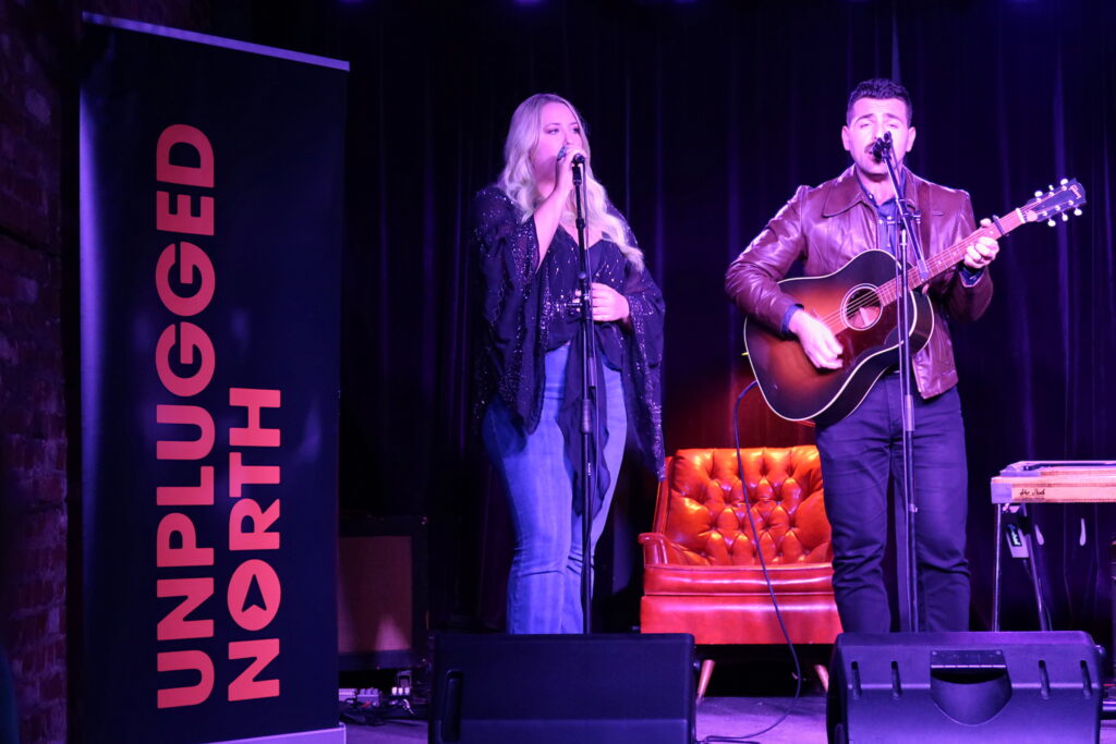 Singers Taylor-Rae (left) and Antonio Larosa (right) performing on a dimly lit stage next to an Unplugged North banner